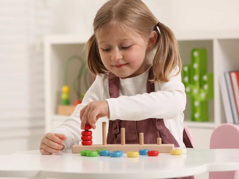 Dievča pri bielom stolíku navlieka farebné drevené krúžky na kolíky počítacej pyramídy.
